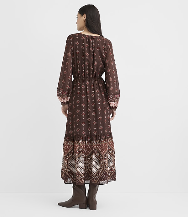 A woman in a long, patterned brown dress and boots stands against a plain white background.