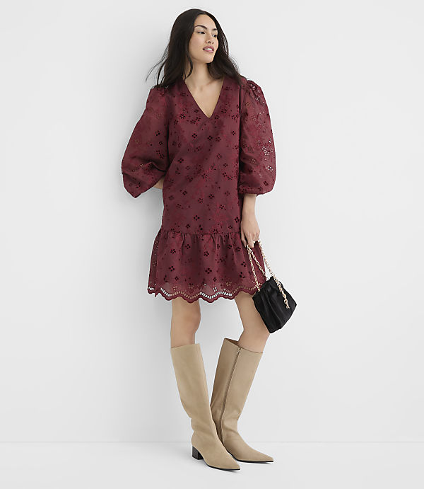 A woman in a burgundy eyelet dress and tan boots poses against a plain white background, holding a black purse.