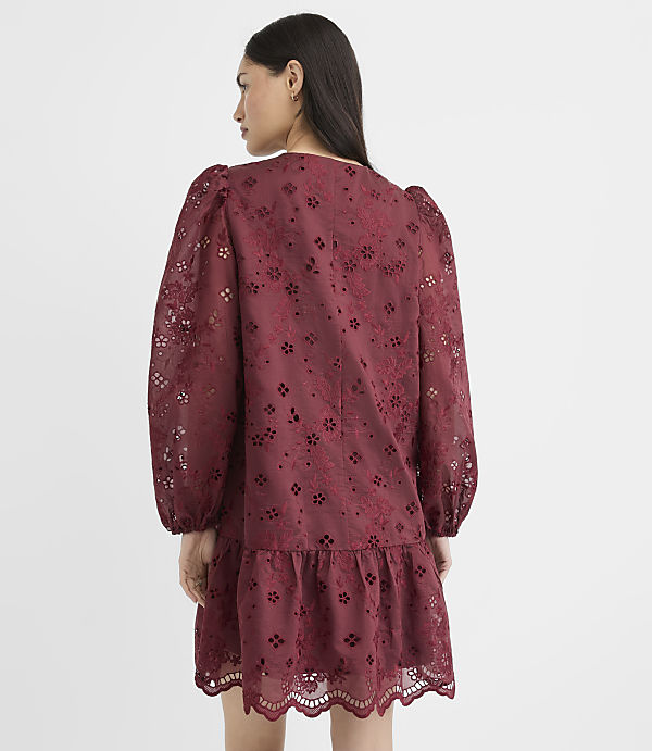 A woman in a maroon eyelet dress with puff sleeves stands against a plain white background.