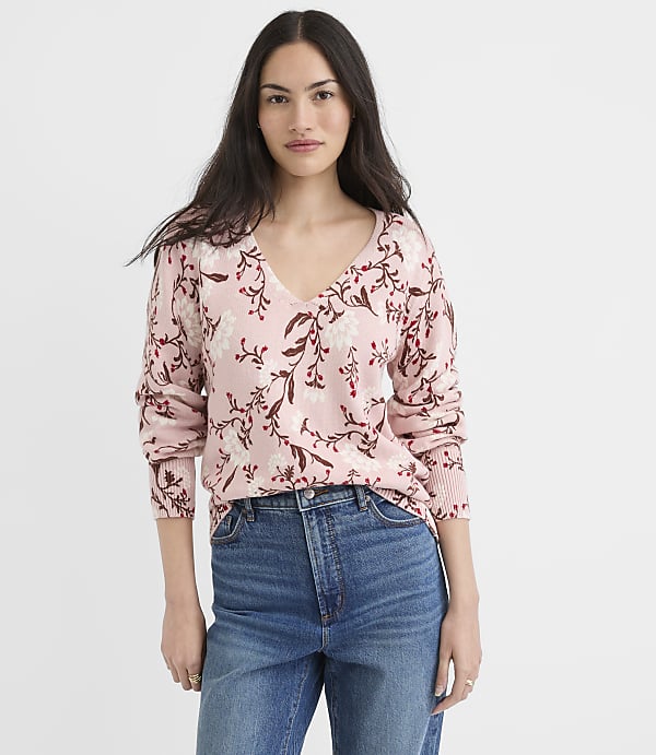 A woman in a floral pink sweater and blue jeans stands against a plain white background.