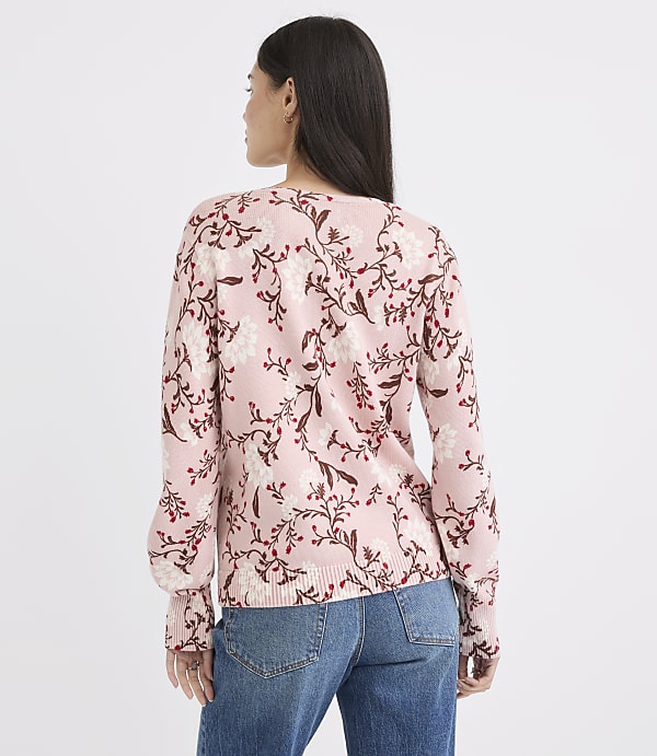 A woman in a pink floral sweater and blue jeans stands against a plain white background.