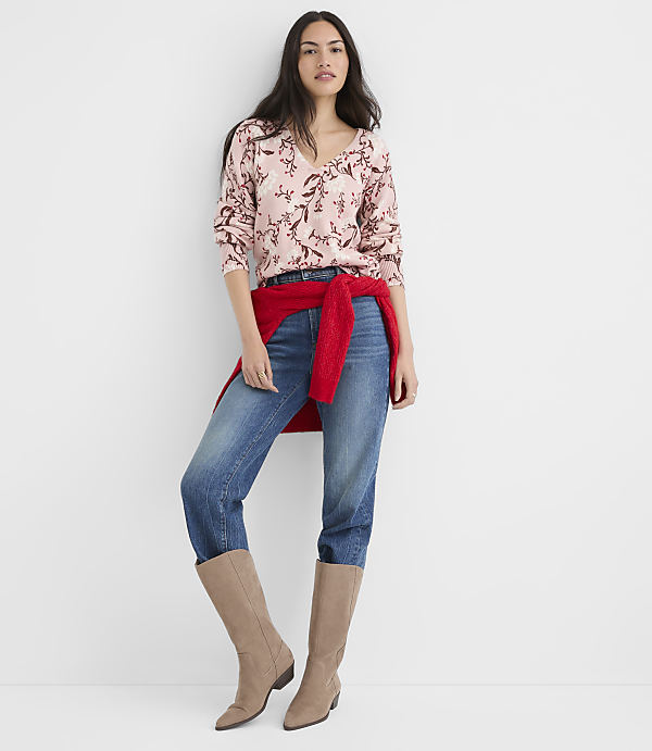 A woman in a floral sweater, jeans, and boots stands against a plain white background.