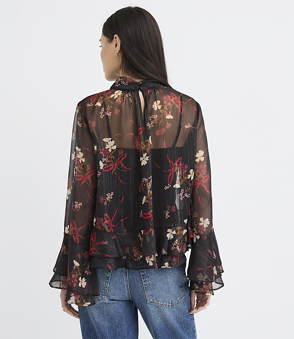 A woman in a sheer floral blouse and jeans stands against a plain white background.