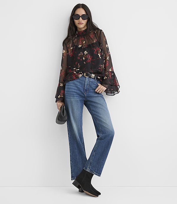 A woman in a floral sheer blouse and high-waisted jeans stands against a plain white background, holding a black clutch.