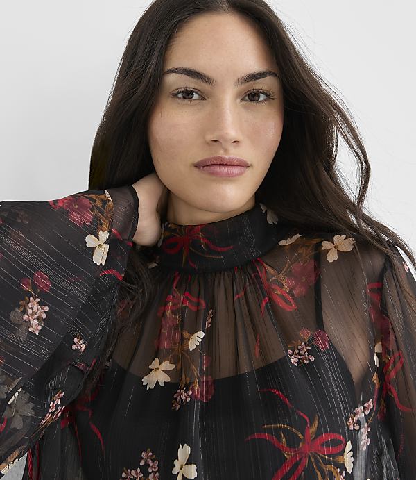 A woman with dark hair and a floral sheer top stands against a plain white background.