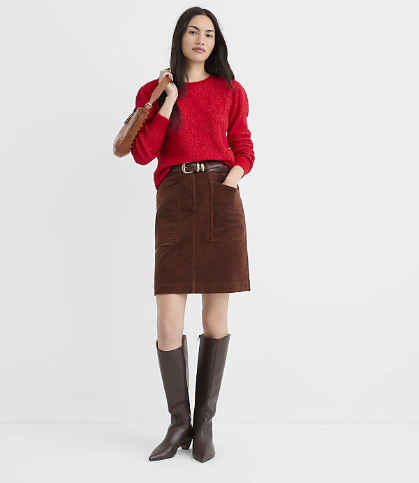 A woman in a red sweater and brown corduroy skirt stands against a plain white background, holding a brown purse over her shoulder.