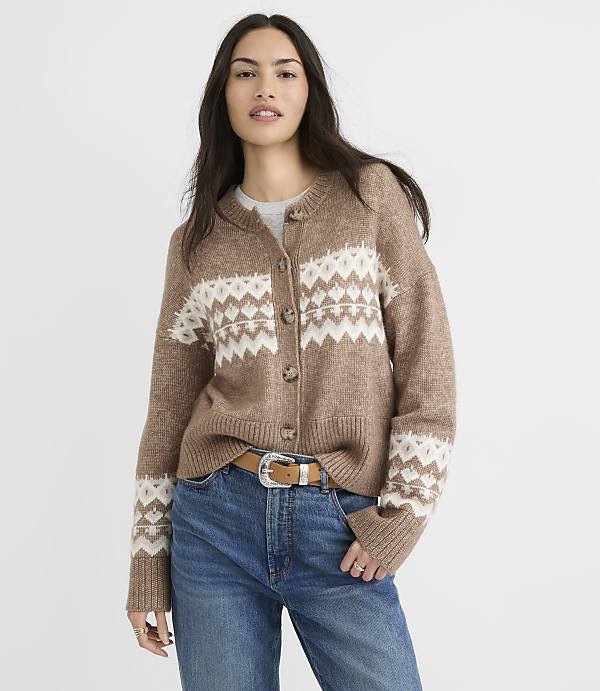 A woman in a brown and white patterned cardigan and blue jeans stands against a plain white background.
