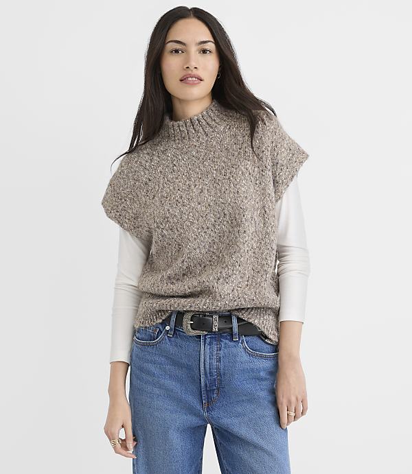 A woman in a taupe knit vest and blue jeans stands against a plain white background.