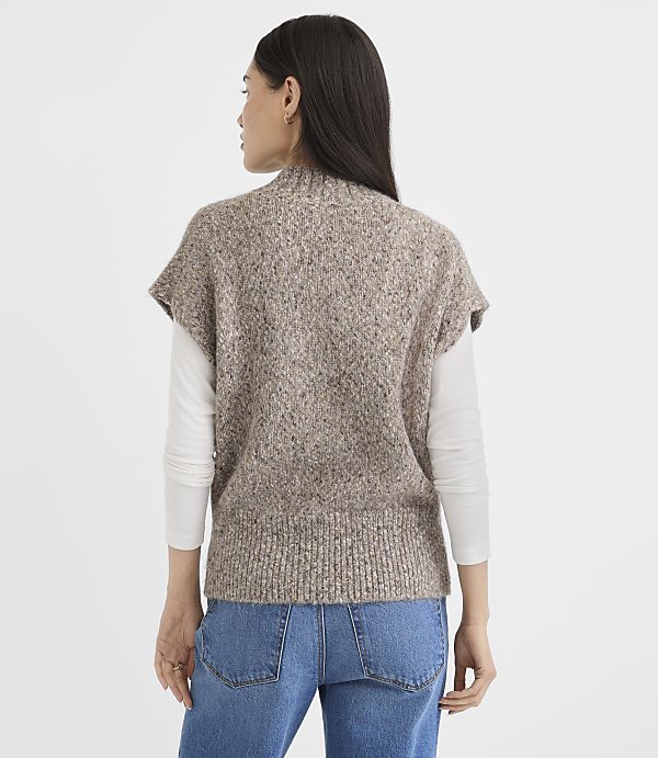 A woman in a textured brown sweater vest and blue jeans stands against a plain white background.