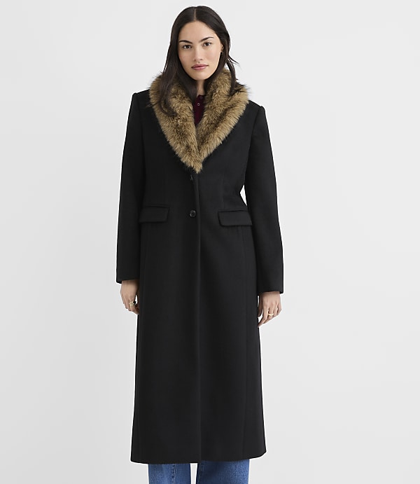 A woman in a long black coat with a fur collar stands against a plain white background.