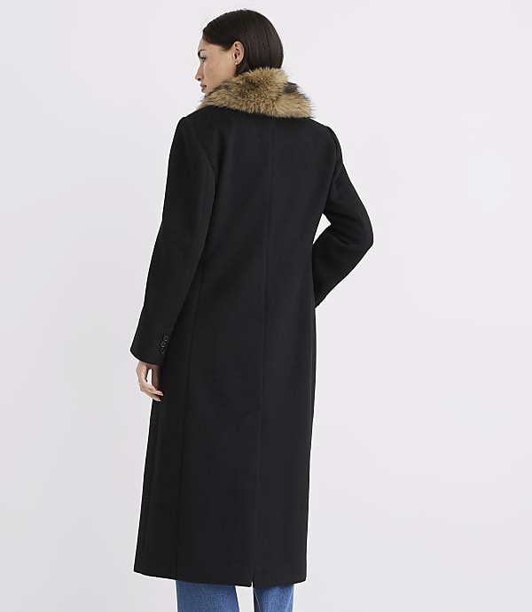 A woman in a long black coat with a fur collar stands against a plain white background.