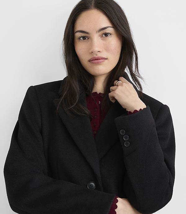 A woman in a black coat and burgundy top stands against a plain white background.