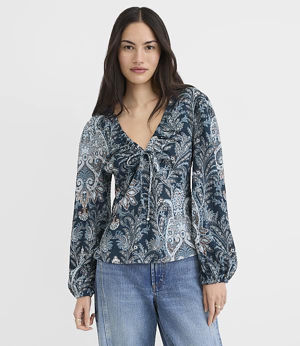 A woman in a patterned teal blouse and blue jeans stands against a plain gray background.