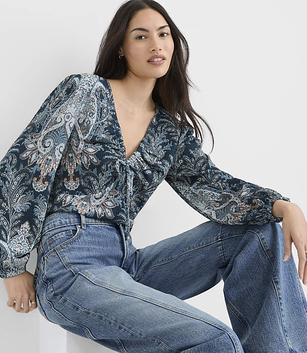 A woman in a patterned blouse and jeans sits against a plain white background.