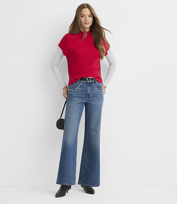 A woman in a red sweater vest and flared jeans stands against a plain white background.
