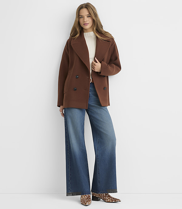 A woman in a brown peacoat and wide-leg jeans stands against a plain white background.