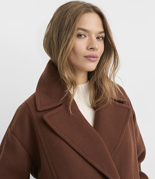 A woman in a brown coat with a white shirt is shown against a plain white background.