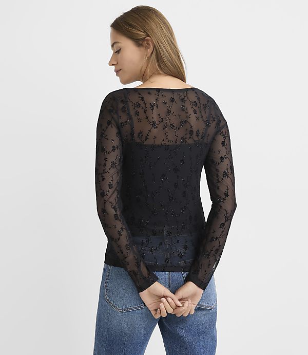 A woman in a black lace top and blue jeans stands against a plain white background.