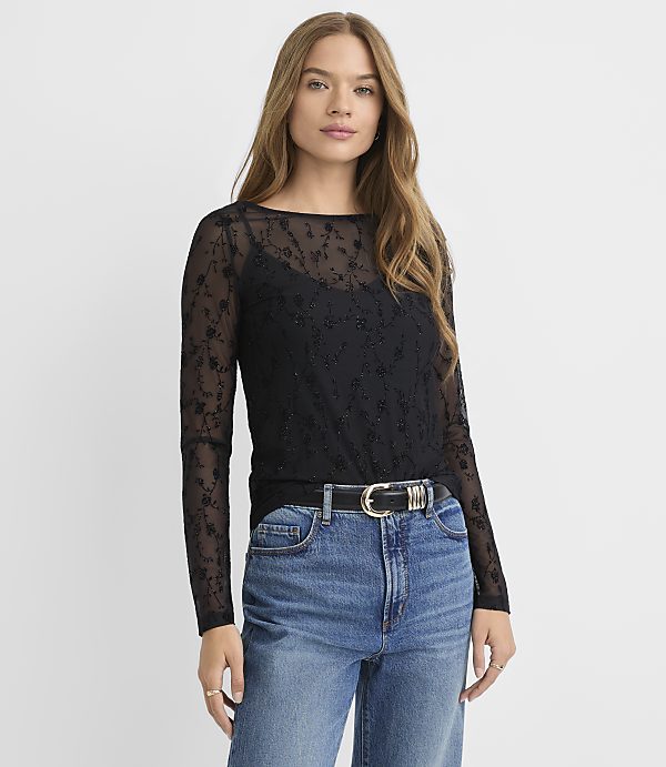 A woman in a black lace top and blue jeans stands against a plain white background.