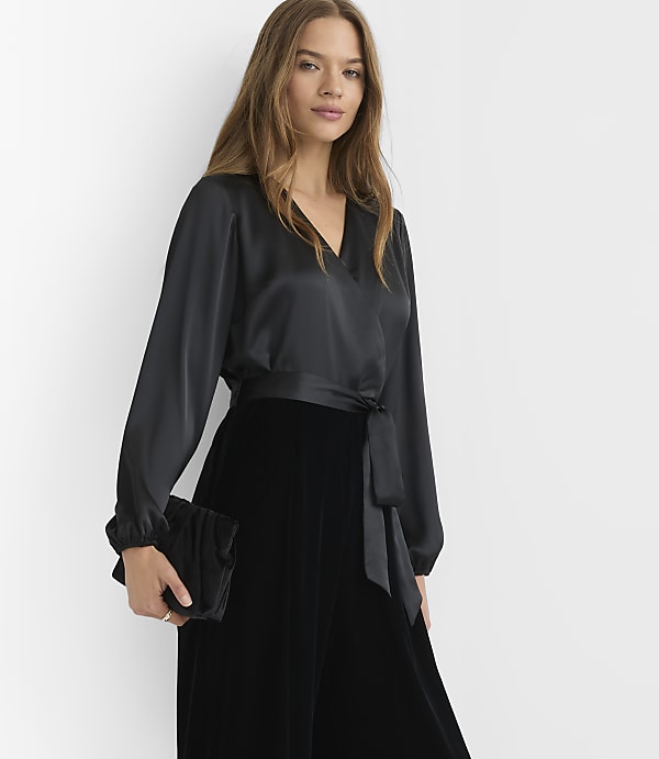 A woman in a black satin wrap top and velvet skirt holds a black clutch against a plain white background.