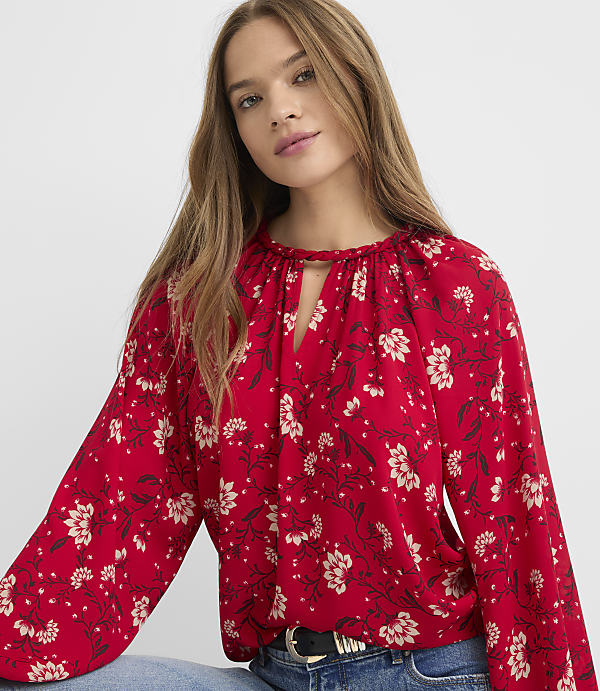 A young woman with long blonde hair wears a red floral blouse with a keyhole neckline, standing against a plain gray background.