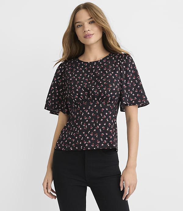 A woman in a black floral top and black pants stands against a plain white background.