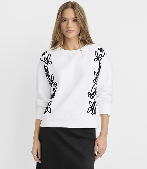 A woman in a white sweatshirt with black floral embroidery stands against a plain white background.