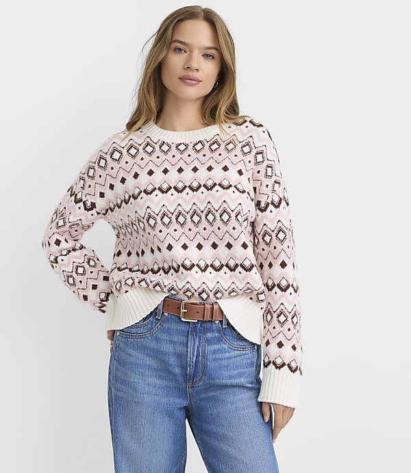 A woman in a patterned sweater and jeans stands against a plain white background.
