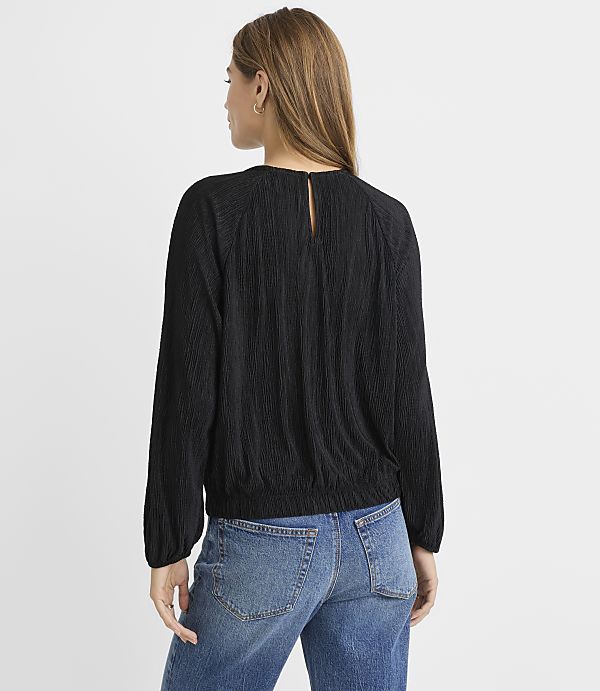 A woman in a black textured blouse and blue jeans stands against a plain gray background.