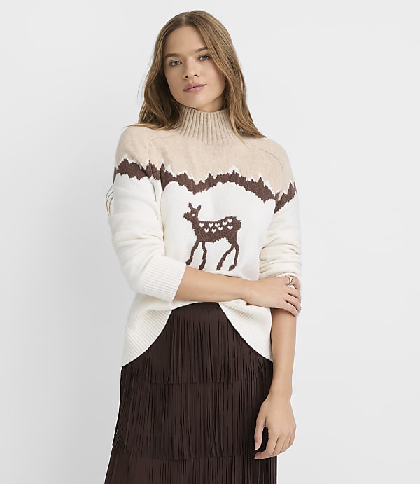 A woman in a cream-colored sweater with a deer and mountain design stands in front of a plain background.