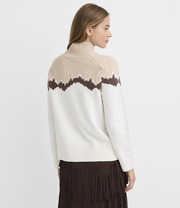 A woman in a cream and tan turtleneck sweater with a brown wavy pattern stands against a plain white background.
