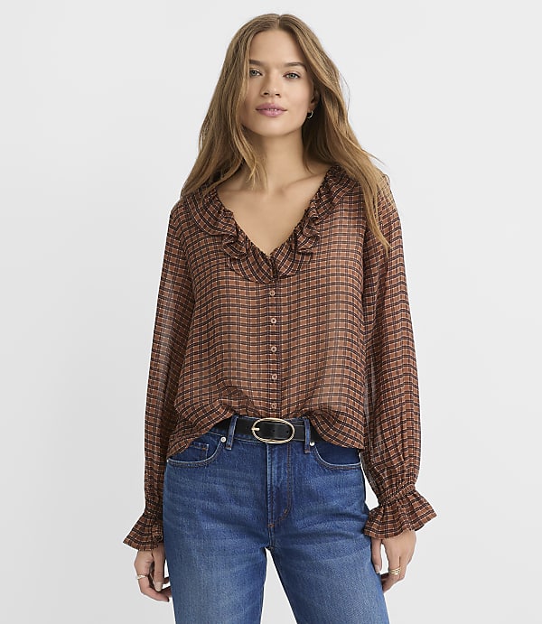 A woman in a brown checkered blouse and blue jeans stands against a plain white background.