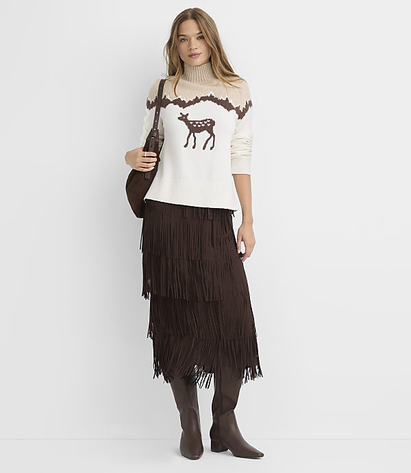 A woman in a cream sweater with a deer motif and a brown fringed skirt stands against a plain white background.