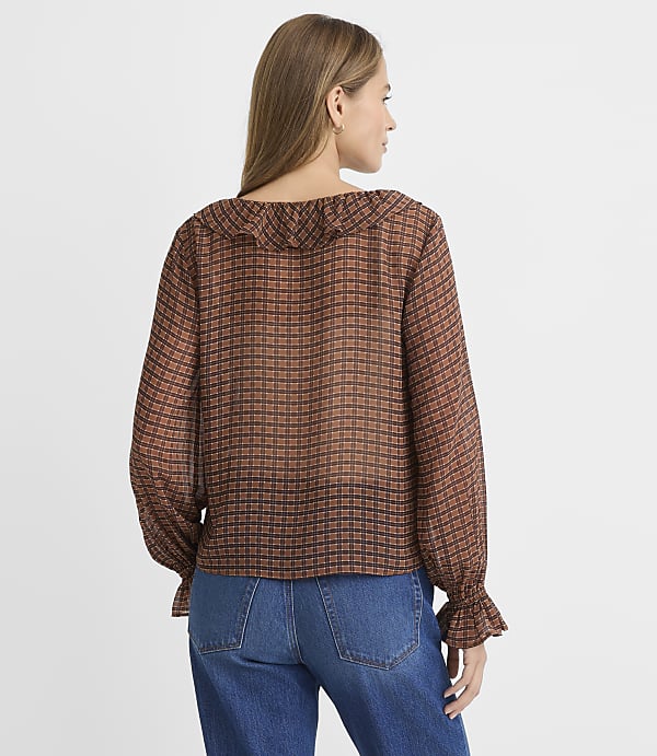 A woman in a brown checkered blouse and blue jeans stands against a plain white background.