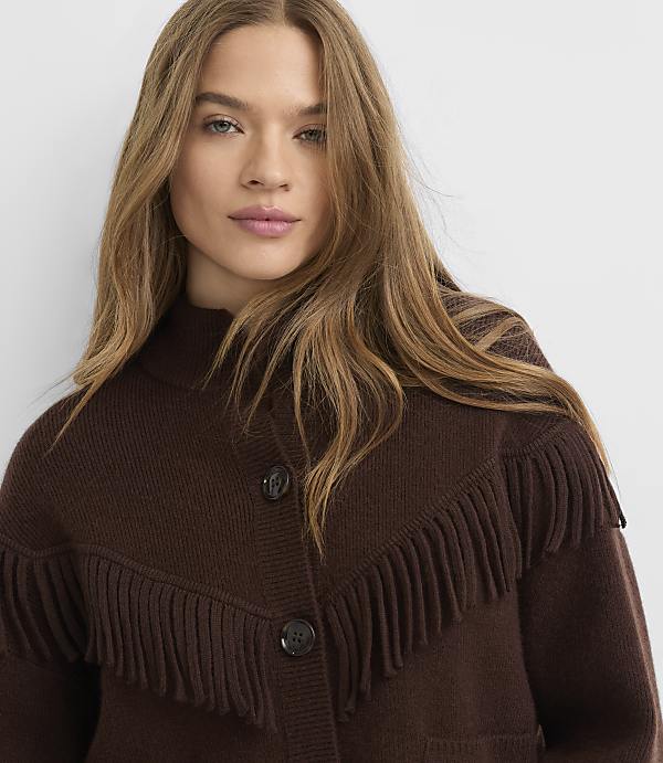 A woman with long, blonde hair wears a brown fringed cardigan against a plain white background.