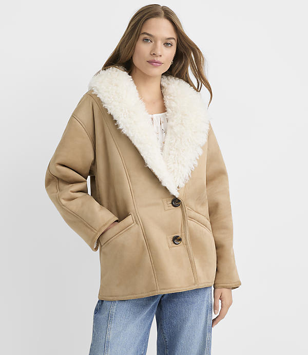 A woman in a tan shearling jacket and blue jeans stands against a plain white background.