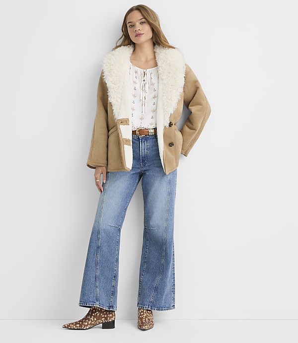 A woman in a shearling-lined jacket and flared jeans stands against a plain white background.