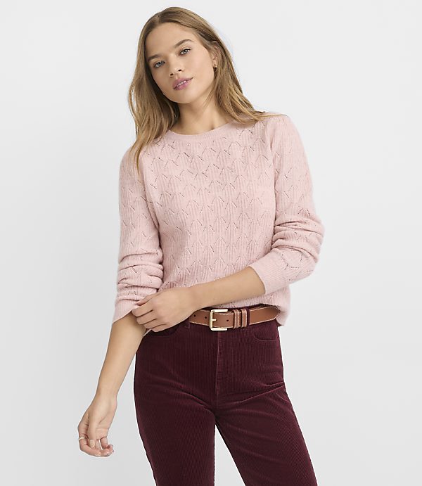 A young woman in a pink sweater and burgundy pants stands against a plain gray background.