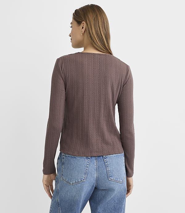 A woman in a brown ribbed long-sleeved shirt and blue jeans stands against a plain, light gray background.