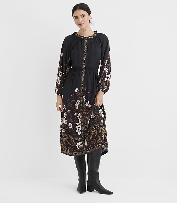A woman in a black floral dress and black boots stands against a plain white background.