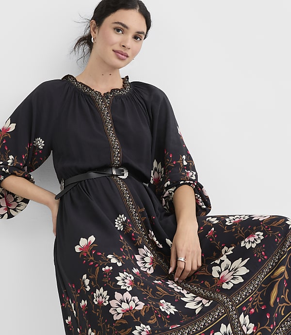 A woman in a black floral dress with a cinched waist stands against a plain white background.