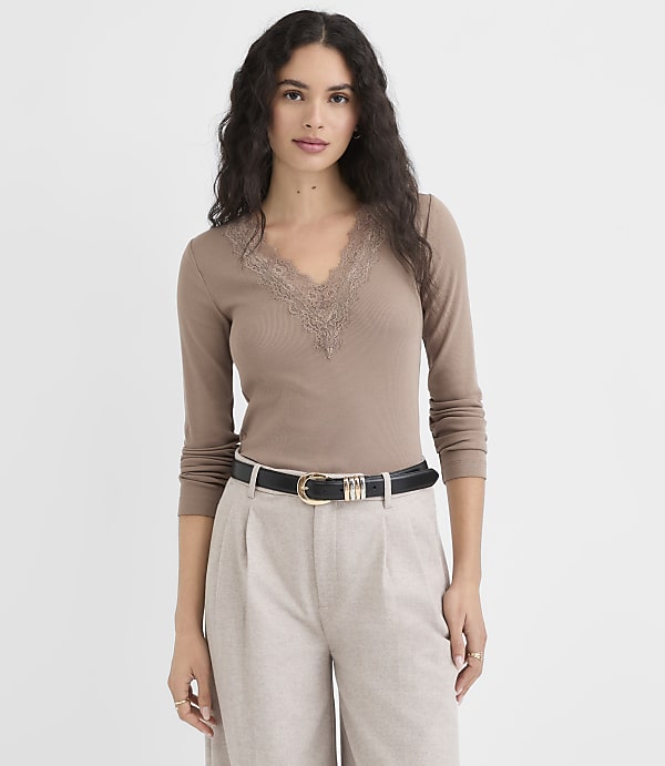 A woman in a brown lace-trimmed top and light-colored pants stands against a plain white background.