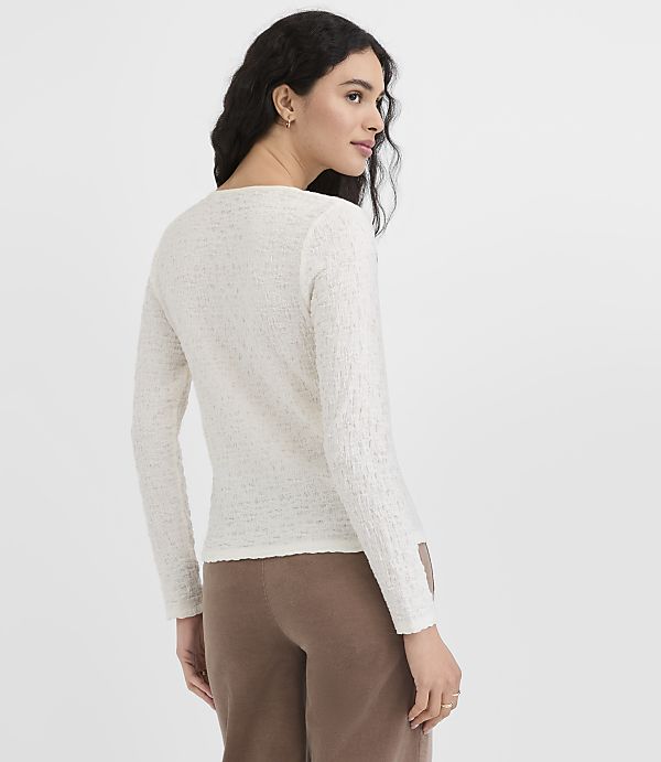 A woman in a cream-colored textured sweater and brown pants stands against a plain white background.