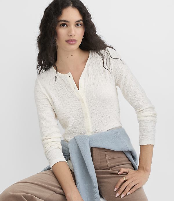 A woman in a cream cardigan and brown pants is seated against a plain white background, with a light blue sweater draped over her shoulders.
