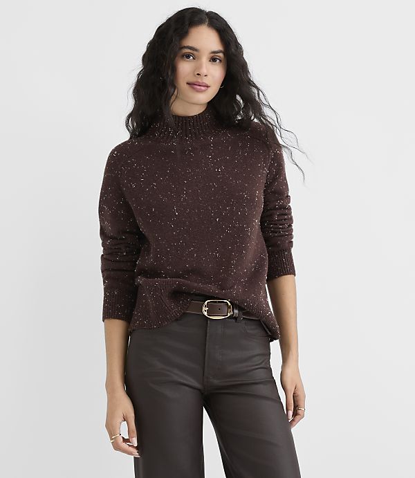 A woman in a speckled brown turtleneck sweater and dark pants stands against a plain gray background.