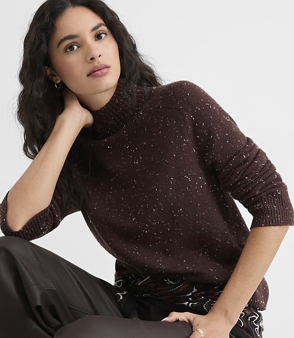 A woman in a speckled brown turtleneck sweater rests her arm on her knee against a plain white background.