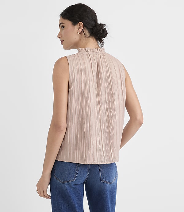 A woman in a textured, light pink sleeveless top and blue jeans stands against a plain, light background.