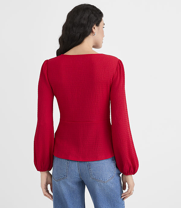 A woman in a red textured top and blue jeans stands against a plain white background.