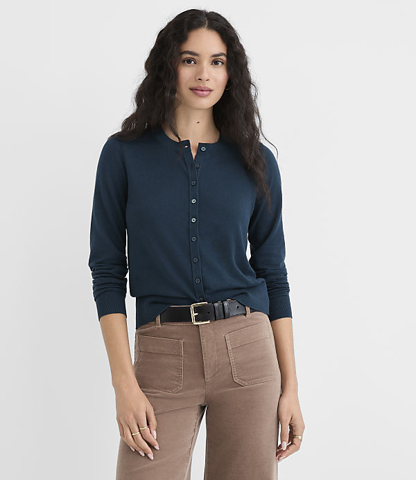 A woman in a teal cardigan and brown pants stands against a plain white background.