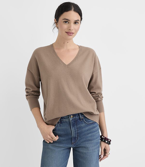 A woman in a brown v-neck sweater and blue jeans stands against a plain white background.